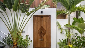 A wooden door with intricate carvings, surrounded by lush tropical plants and a small garden area with stepping stones.