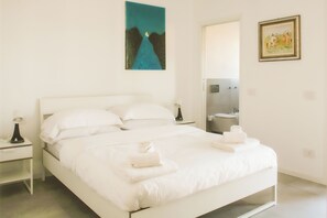 A white bed with pillows and folded towels, a nightstand with a lamp, a painting on the wall, and a bathroom visible through an open door. - BnButler - Govone 78 (Milan)