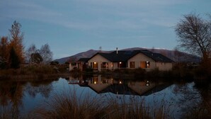 Ein zweistöckiges Haus mit einem spiegelnden Teich davor, umgeben von Bäumen und Hügeln.