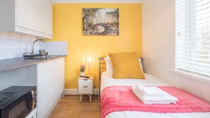 A compact hotel room with a kitchenette, a bed with a pink blanket, a yellow pillow, and a painting of a bridge on the wall. - The Modern Cambridge Studio 3 (Cambridge)