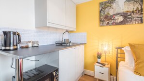 A compact kitchen with white cabinets, a stainless steel kettle, a coffee maker, and a sink. There is a small table with a lamp and a plant. A bed with a yellow pillow is visible in the corner. - The Modern Cambridge Studio 3 (Cambridge)