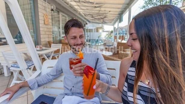Deux personnes sont assises à une table à l’extérieur, devant des boissons, sur une terrasse couverte, avec vue sur la rue.