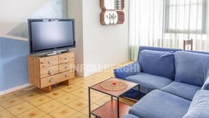 Un salon avec un canapé bleu, un meuble TV en bois sur lequel est posée une télévision à écran plat, une petite table avec un set de table, et une étagère murale décorée de divers objets.