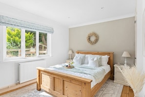 A bedroom with a wooden bed, a radiator, a window with a view of greenery, and a wall-mounted wreath. - Bramblewood (Maidencombe)