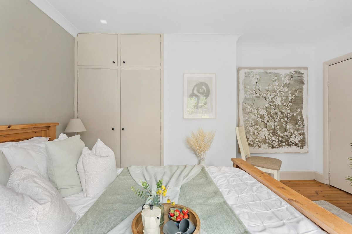 A bedroom with a bed, a bedside table with a vase and fruit, a chair, and a large abstract painting on the wall.