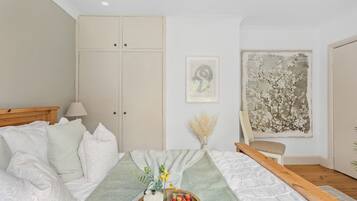 A bedroom with a bed, a bedside table with a vase and fruit, a chair, and a large abstract painting on the wall.