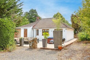 A single-story house with a tiled roof, a front porch, and a garden area with potted plants and a barbecue grill. - Bramblewood (Maidencombe)