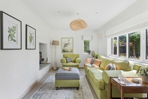 A living room with a green sofa, a matching ottoman, and a coffee table. There are framed botanical prints on the wall, a lamp, and a large window with a view of greenery outside. - Bramblewood (Maidencombe)