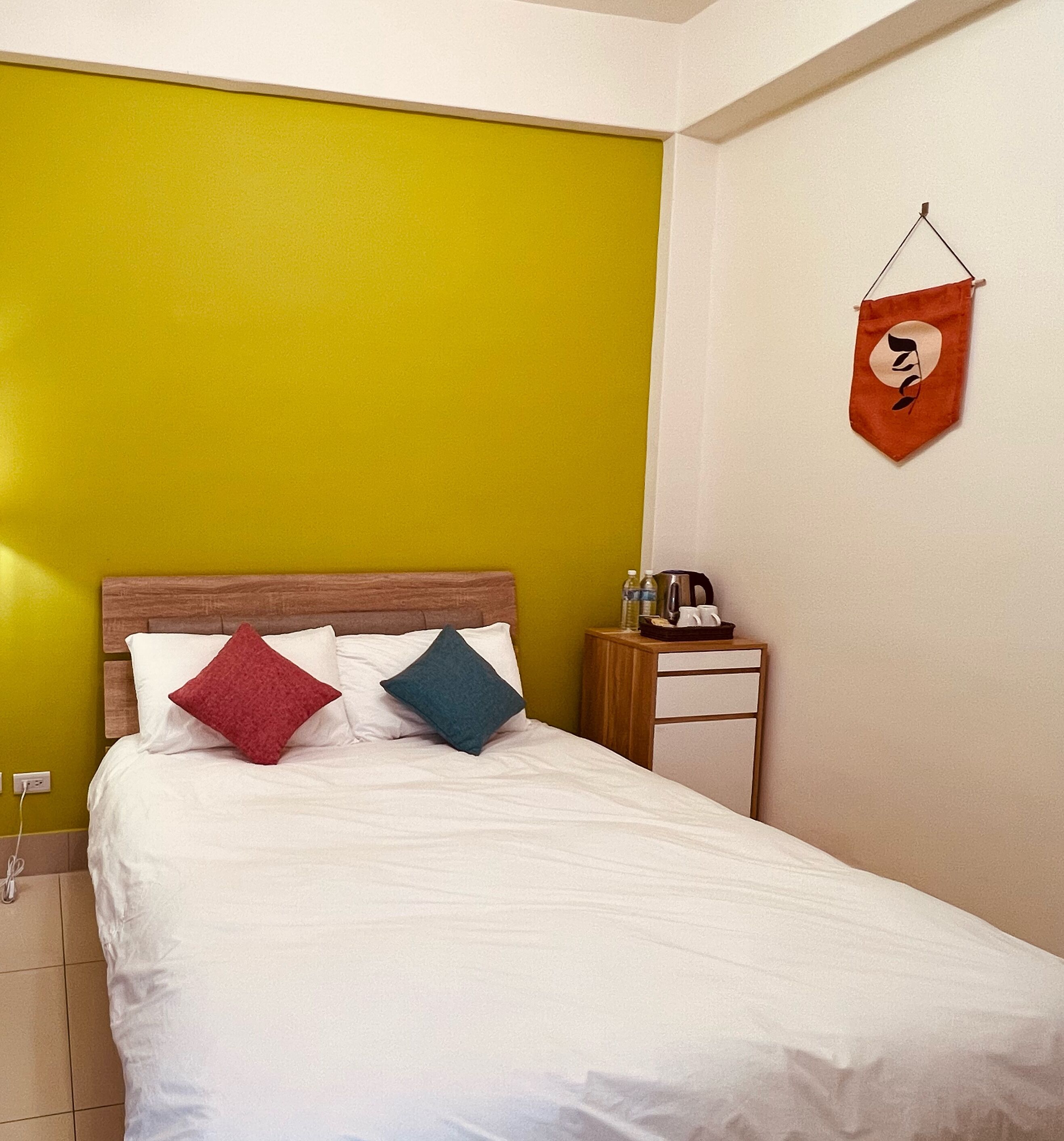 Habitación de hotel con cama, mesita de noche, bolso colgado en la pared y pared amarilla.