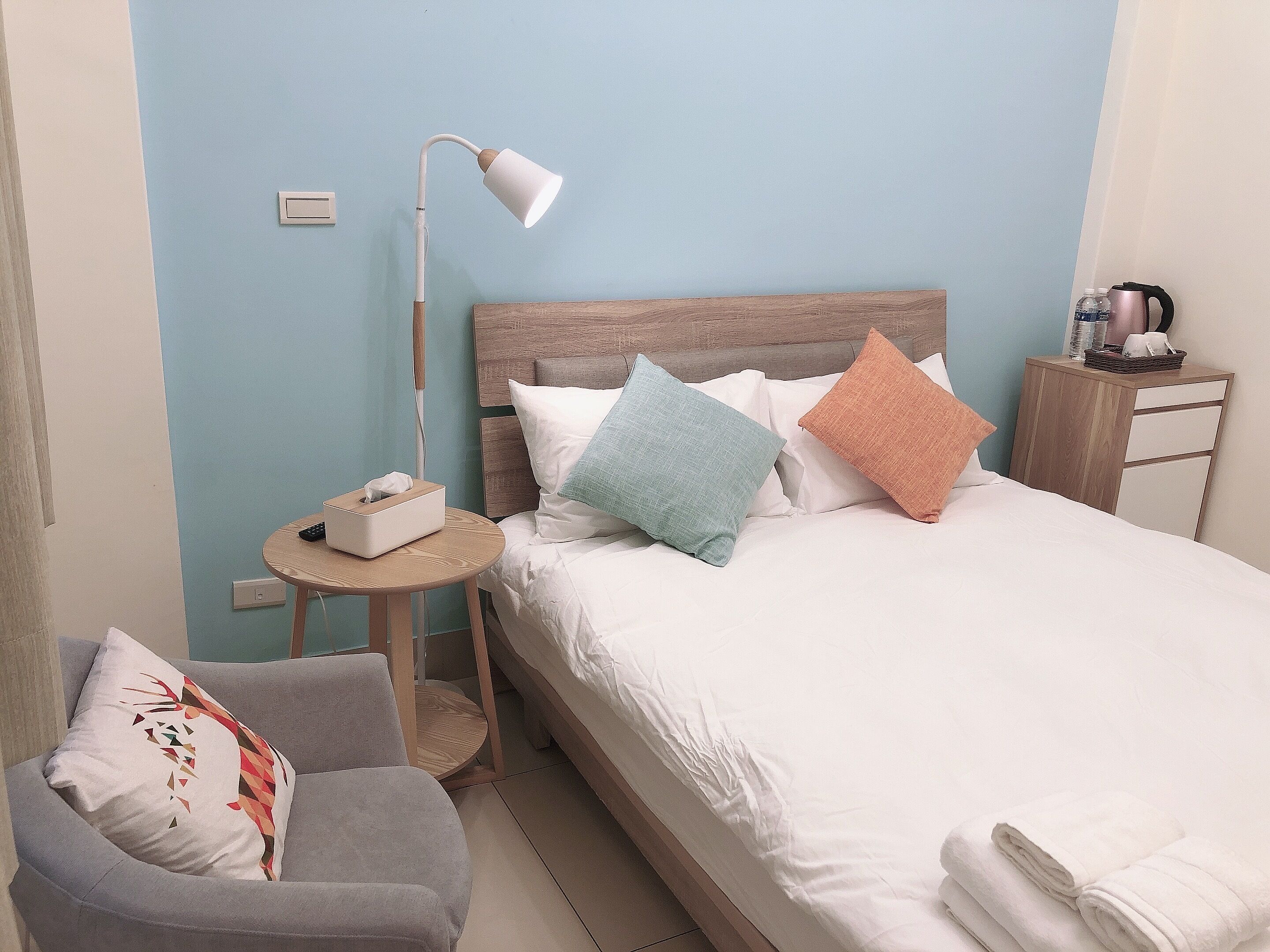 Habitación de hotel ordenada con cama, mesita de noche, lámpara, silla y una pequeña mesa con caja de pañuelos.