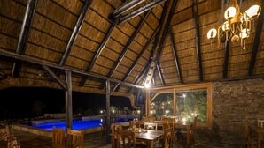 Un espacio de comedor al aire libre con mesas y sillas de madera, techo de paja y una piscina al fondo.