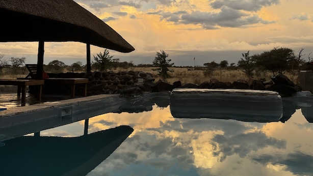 Un área de piscina tranquila al aire libre con una estructura de techo de paja, rodeada de vegetación natural y cielos despejados.