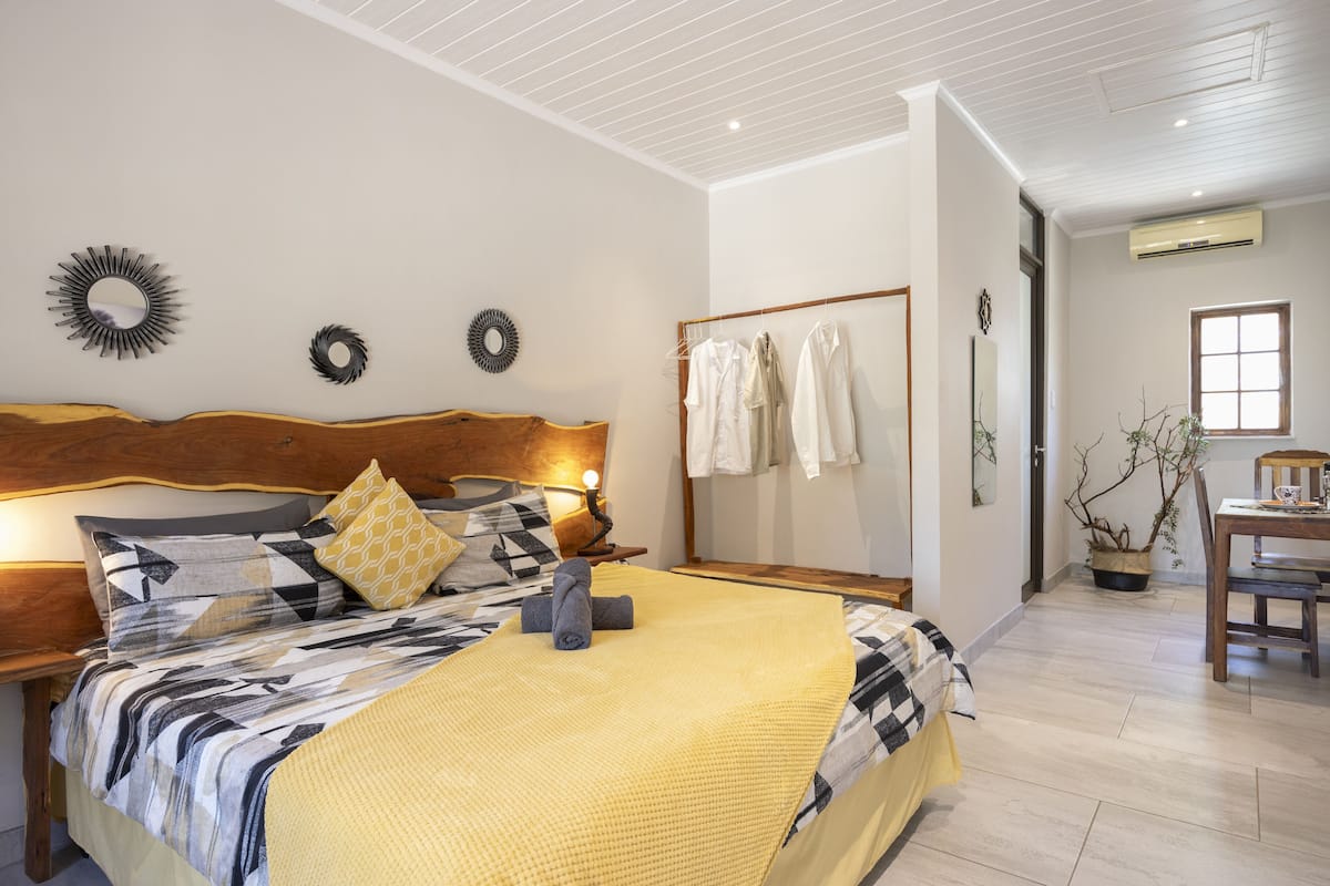 A bedroom with a wooden headboard, a bed with a patterned bedspread, a bedside table with a lamp, and a wall-mounted mirror.