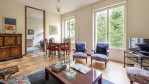A spacious living room with a wooden floor, a dining area with red chairs, two floral armchairs, a coffee table with books, and large windows offering a view of greenery. - Milano Classy Heritage Home: quiet and prestigious apartment CITY CENTER - 4 BDR (Milano)