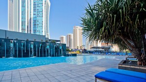 Un área de piscina con sillas reclinables azules, una fuente de agua y edificios altos al fondo.