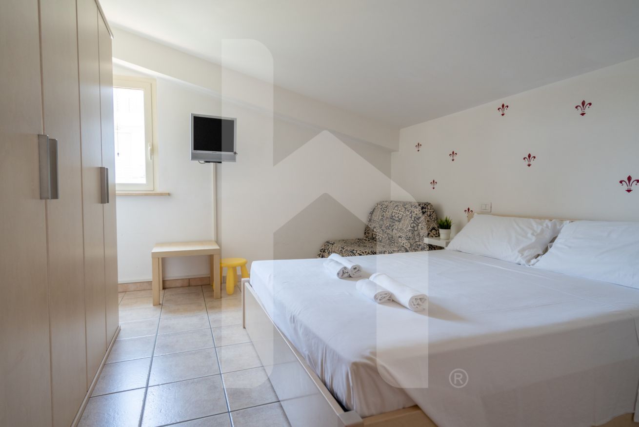Un dormitorio con una cama, un televisor montado en la pared, un pequeño banco y una ventana.