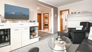 A modern living room with a flat-screen TV, a glass coffee table, a black leather sofa, and a kitchen area with wooden cabinets. - Wohnung Wesernest in Büsum (Büsum)