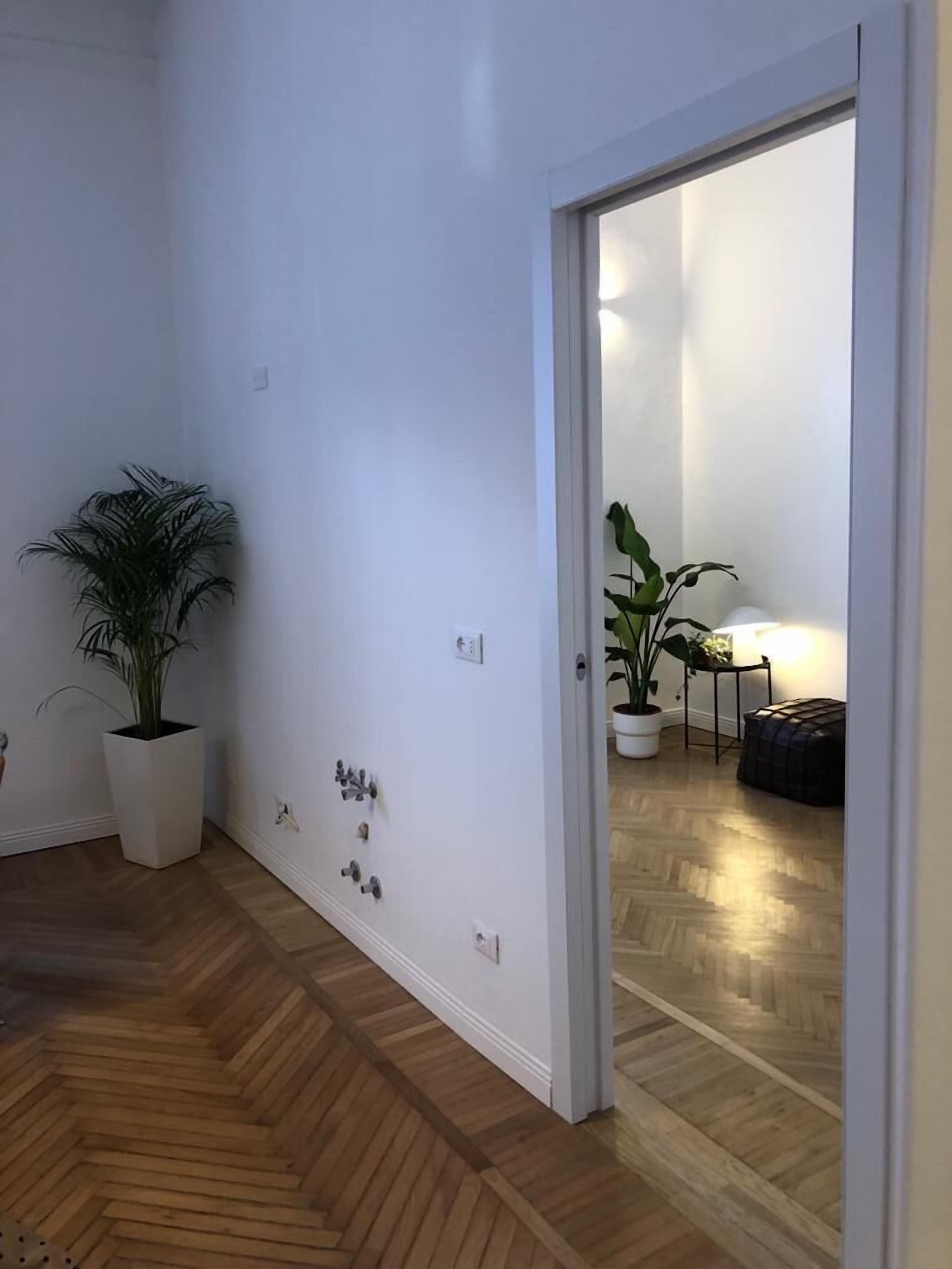 A room with a wooden herringbone floor, white walls, and a door leading to another room. There are two potted plants, one on the left and one on the right.