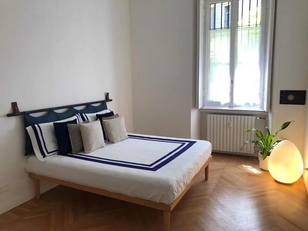 Un dormitorio con cama, radiador, ventana con persianas y lámpara de noche.