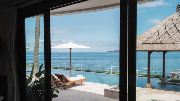 A view from a room with a large window overlooking a pool area with lounge chairs and a thatched-roof structure. - Villa Blanca by Alfred in Bali (Manggis)