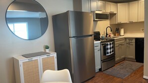 Una cocina compacta equipada con refrigerador, microondas, estufa y lavavajillas. Se observa un espejo redondo en la pared, una pequeña planta y una toalla de cuadros colgada del horno.