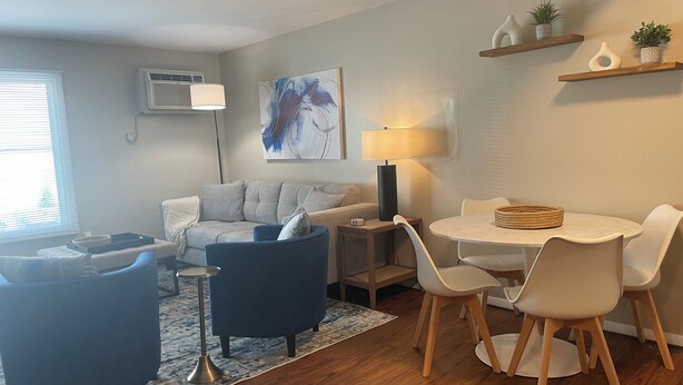 Una sala de estar moderna con un sofá, un sillón azul, una mesa de comedor con sillas, una lámpara y un cuadro en la pared.