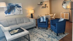 Una sala de estar moderna con un sofá gris, sillones azules, una mesa de centro y un tapete estampado. Se observa un cuadro en la pared, una lámpara y una pequeña mesa con sillas.