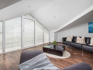 A modern loft apartment with a sloped ceiling, large floor-to-ceiling windows, a black leather sofa, and a glass coffee table.