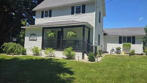 A two-story house with a porch, a small garden, and a clear blue sky. - Cozy Farmhouse w/ short walk to a private  lake
 (Delton)