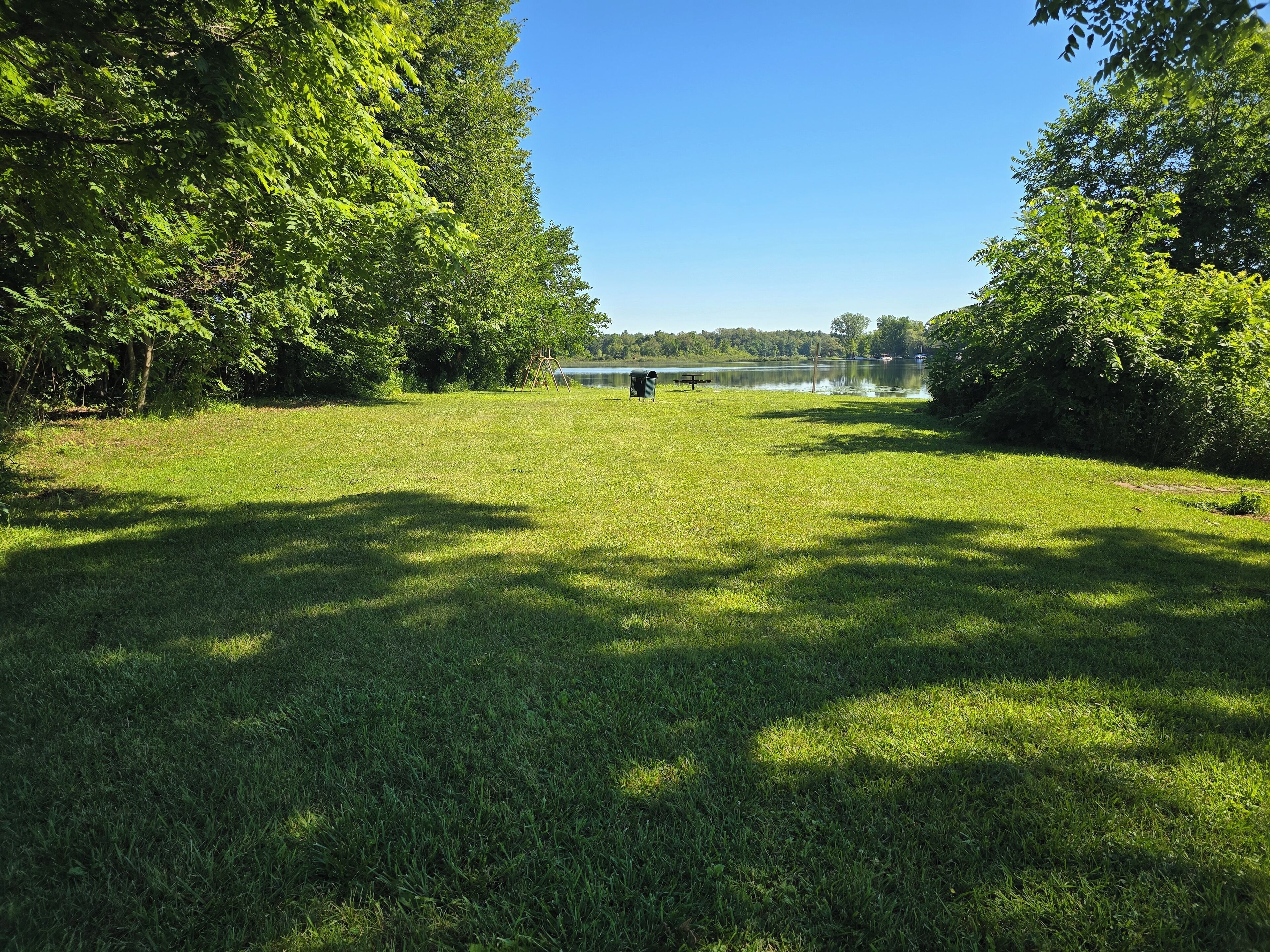 Un campo cubierto de césped con un conjunto de columpios y una mesa de picnic a lo lejos, rodeado de árboles y un lago.