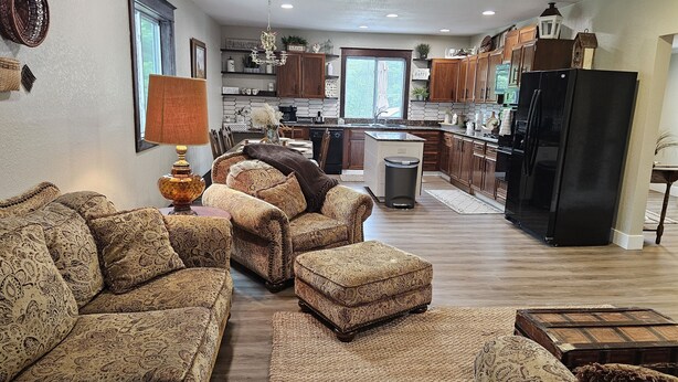 A spacious living room with a sofa set, a coffee table, and a lamp. The kitchen is visible in the background with wooden cabinets and a black refrigerator. - Cozy Farmhouse w/ short walk to a private  lake
 (Delton)