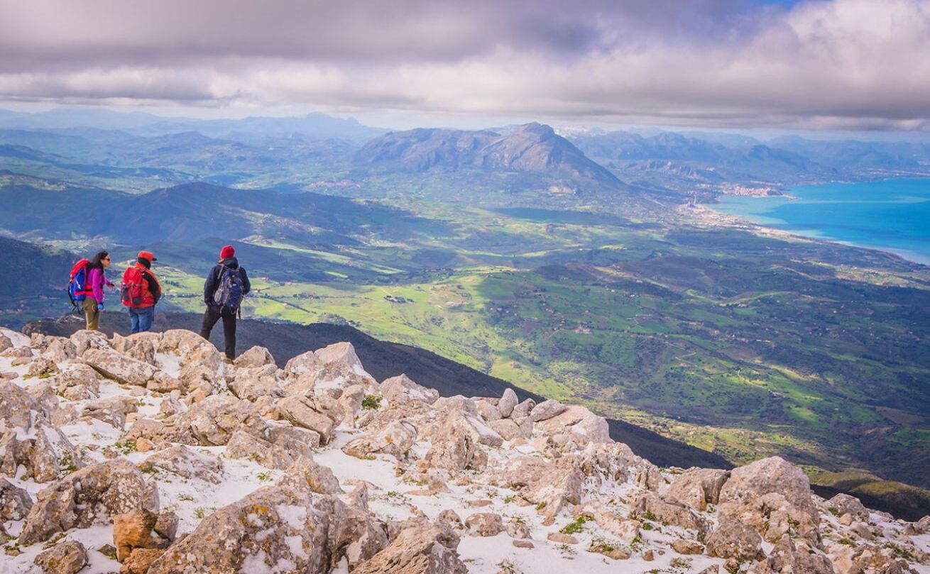 A group of hikers on a rocky mountain peak with a view of a valley and a body of water below.