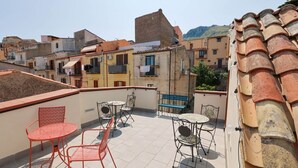 Une terrasse sur le toit, dotée d’un espace extérieur avec des tables et des chaises, offrant une vue sur un quartier résidentiel aux toits de tuiles et sur une montagne en arrière-plan.