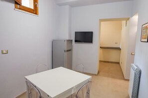 Et lille, kompakt værelse med et hvidt bord og stole, et indbygget køleskab, et tv ophængt på væggen og en radiator.