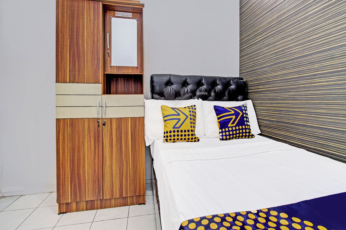A bedroom with a wooden wardrobe, a bed with white bedding and patterned pillows, and a dark upholstered headboard.