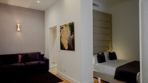 A modern hotel room with a dark-colored sofa, a bed with a headboard, and a painting on the wall. - Màs Boutique Hotel (Rome)