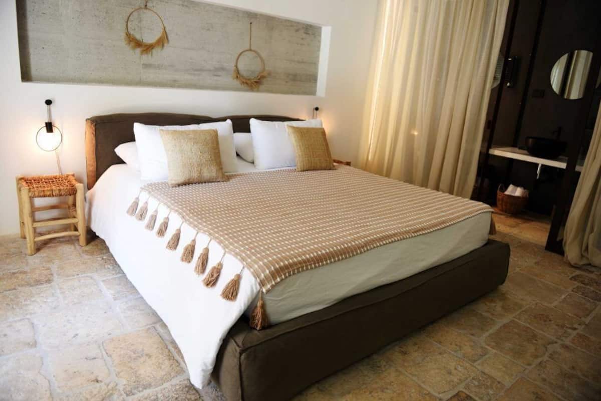 A bedroom with a large bed, a headboard, a nightstand, a wall-mounted artwork, and a bathroom with a mirror and sink.