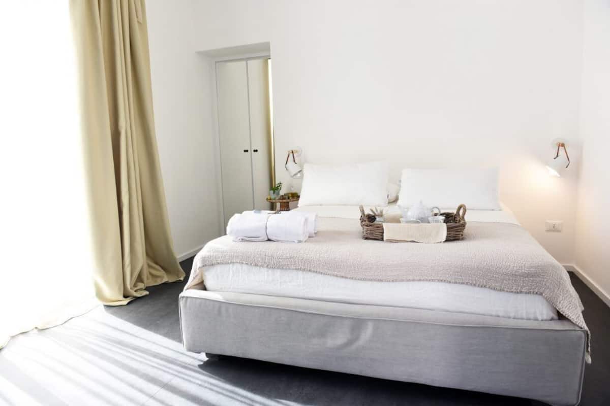 A neatly made bed with white linens, a grey mattress, and a light-colored blanket. There are two white pillows and a small basket with towels on the bed.