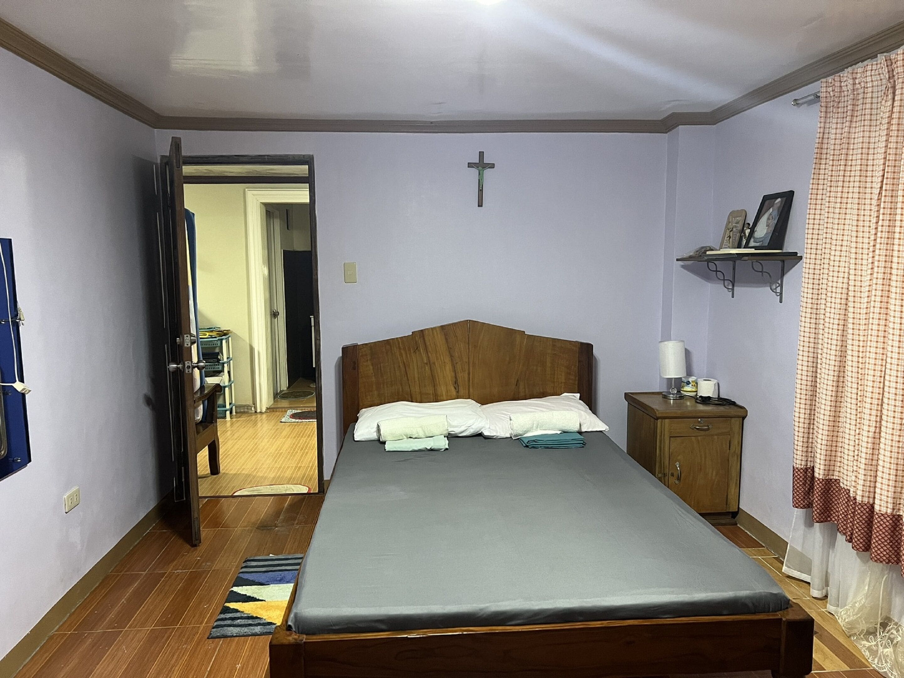 Una camera da letto con un letto di legno, un comodino con una lampada, una croce appesa al muro e una mensola con un quadro.