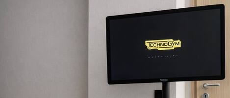 A hotel room with a black TV on a stand, a black speaker, and a black bag with 'TECHNOGYM' on it.