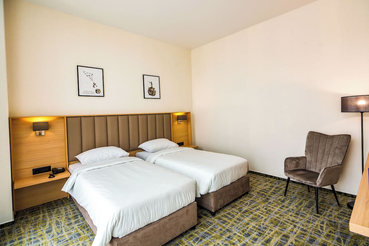 A hotel room with two beds, a chair, a lamp, and artwork on the wall.