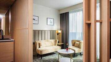 A hotel room with a sofa, armchair, and a small round table with a bottle. There is a wooden partition on one side and a window with curtains.