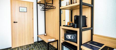 A compact room with a wooden door, a small desk with a chair, a mini-fridge, a coffee machine, and a shelf with various items.