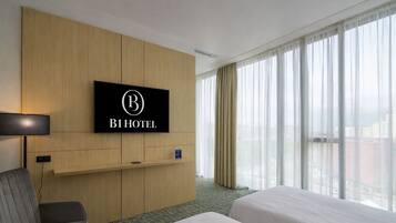 A hotel room with a bed, a TV mounted on a wooden panel, a desk with a chair, and a large window with curtains.