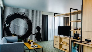 A modern hotel room with a large black artwork, a flat-screen TV, a sofa, a coffee table, and a wooden cabinet with shelves.