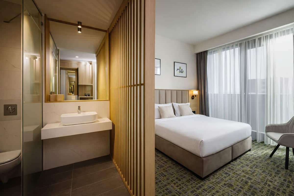 A hotel room with a bathroom, a bed, a chair, and a large window.