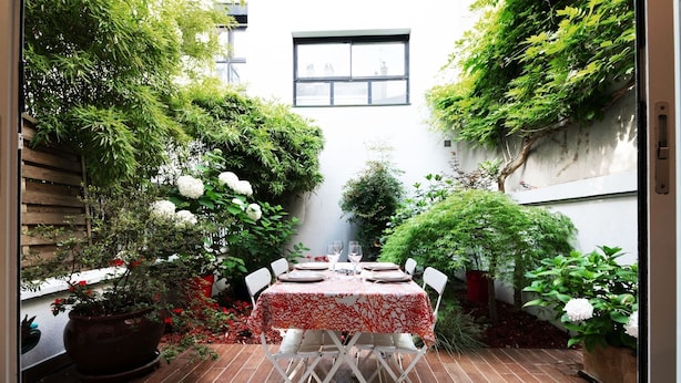 An outdoor dining area with a table set for two, surrounded by potted plants and a white wall.
