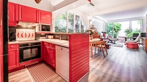 A modern kitchen with red cabinets, a dishwasher, and a microwave. There is a dining area with a table and chairs, and a large window offering a view of greenery.
