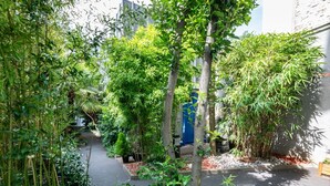 A narrow pathway flanked by dense greenery and bamboo plants, leading to a blue door.