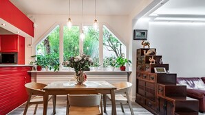A dining area with a wooden table and chairs, a red kitchen area, and a view of greenery through large windows.